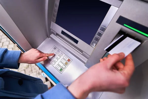 Közeli kép egy bankautomatáról, amelyen valaki gombokat nyom és bankkártyát tart a terminál mellett.