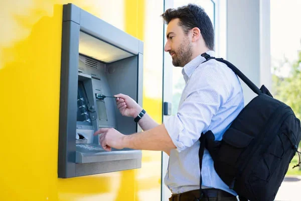 Egy férfi bankautomata előtt áll, és a gépet használja, hátizsákkal a vállán.